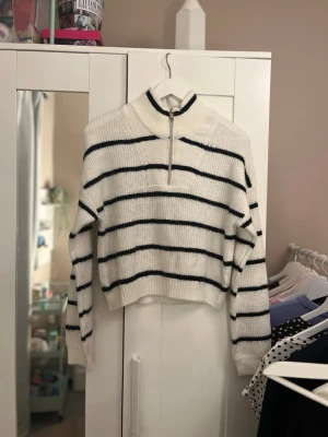 Vit stickad tröja med svarta ränder - Vit stickad tröja med svarta horisontella ränder och halv dragkedja framtill. Tröjan har hög krage, långa ärmar och ribbade muddar. Croppad modell som ger en trendig look. Perfekt för lager på lager och enkel att styla till jeans eller kjol.