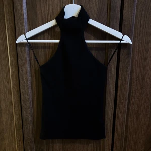 Svart halterneck topp med polokrage - Snygg svart halterneck topp med hög polokrage. Toppen har en tight passform och är gjord i ett stretchigt material som sitter skönt mot kroppen. Perfekt för att skapa en trendig och stilren look.