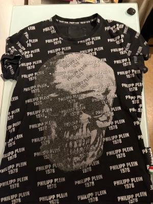 Svart Philipp Plein t-shirt med dödskalle - Cool svart t-shirt från Philipp Plein med stort dödskalletryck framtill och all-over logotyptext 'PHILIPP PLEIN 1978' i vitt. T-shirten har rund hals och korta ärmar, perfekt för dig som gillar edgy streetwear. Materialet är mjukt och skönt bomull.