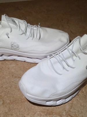 Vita Loewe x On Cloudtilt sneakers - Säljer ett par helvita Loewe x On Cloudtilt sneakers med minimalistisk design och vågig sula. Skorna har tunna elastiska snören, diskret Loewe-logga på sidan och är tillverkade i mesh och syntetmaterial för en sportig look. SNABB AFFÄR 500KR