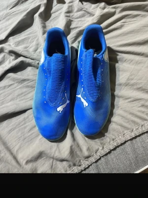 Blåa Puma fotbollsskor - Säljer ett par blåa fotbollsskor från Puma med vit logga på ovansidan. Skorna har en strumpliknande passform och mönstrad sula i ljusgrönt gummi, perfekt för spel på konstgräs eller inomhusplan. Snygg design med diskreta vita detaljer och modern look.