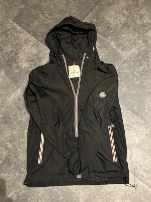 Svart Moncler vindjacka med huva - Svart vindjacka från Moncler med huva och dragkedja. Jackan har vita och röda ränder längs dragkedjor och ärmslut samt Moncler-logga på bröstet. Tillverkad i lätt material perfekt för blåsiga dagar. Snygg lite äldre modell det är därför priset är som det är. Helt äkta. Storlek 2 vilket är small.