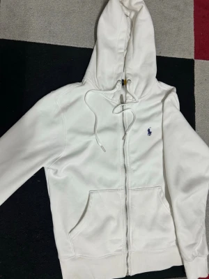 Vit hoodie från Polo Ralph Lauren - Säljer en clean vit hoodie från Polo Ralph Lauren med dragkedja, huva med dragsko och kängurufickor. Klassisk broderad logga i mörkblått på bröstet. Tillverkad i mjuk bomullsmix som är skön att ha på sig. Perfekt för en chill och stilren look.