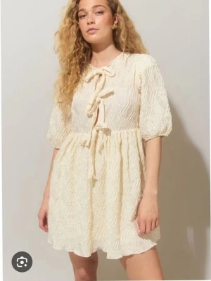 Kort ljus klänning med knyt - Supersöt kort klänning i ljus beige med strukturmönster och puffärmar. Klänningen har flera knyt framtill och en luftig, flowy passform. Perfekt för varma dagar och ger en romantisk vibe.