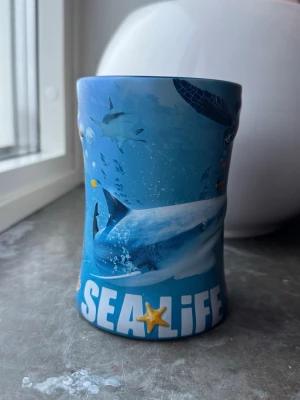 Blå SEA LIFE mugg med havsmotiv - Cool mugg i blått med SEA LIFE-tryck och motiv av haj, sköldpadda och färgglada fiskar. Muggen har en unik form och ett stort handtag, perfekt för dig som gillar havet och marina djur. Tillverkad i keramik och dekorerad med undervattensscener. 