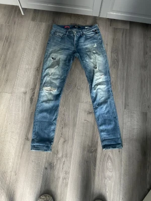 Blåa replay slitna jeans med hål  - Ett par blå jeans med skinny fit och slitningar på både fram- och baksida. Jeansen har flera hål och slitna detaljer på låren och knäna för en trendig, avslappnad look. Klassisk femficksmodell med dragkedja och knapp. Perfekta för dig som gillar en edgy stil. 