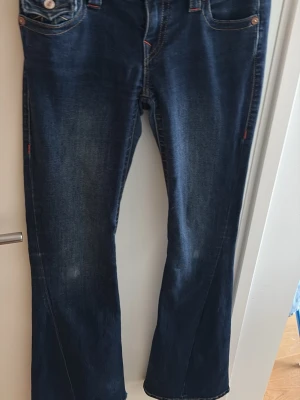 Mörkblå bootcut jeans med kontrastsömmar - Snygga mörkblå bootcut jeans med kontrastsömmar och klassisk femficksdesign. Jeansen har lätt slitna detaljer framtill och orangea sömmar vid fickorna. Perfekta för en avslappnad och trendig look. Upptrampade längst ner.