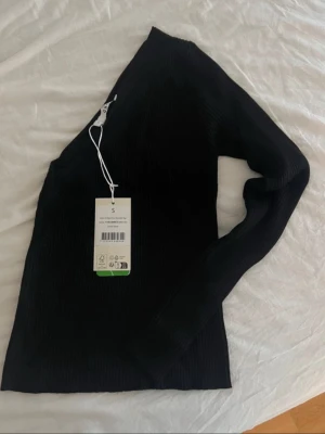 Svart oneshoulder ribbad topp - Trendig svart oneshoulder topp med lång ärm och ribbad struktur. Perfekt för dig som vill ha en stilren och modern look. Toppen är figurnära och har en unik asymmetrisk design som sticker ut.
