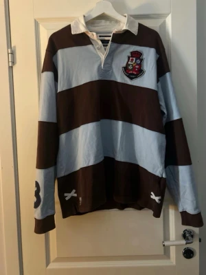 Hackett London skjorta  - Säljer en långärmad rugbytröja med breda bruna och ljusblå ränder, vitt krage och klubbmärke på bröstet. Stor siffra 3 på ryggen och ärmen. Klassisk rugbylook med knappar i halsen och robust bomullsmaterial.