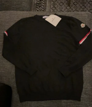 Moncler tröja - Svart långärmad tröja från Moncler med klassisk rund hals och ribbade muddar. På ärmen finns en röd och vit rand samt Moncler-logga. Tröjan har en clean look och är gjord i mjuk bomull som känns skön mot huden. Perfekt för dig som gillar stilrena märkesplagg. Helt ny med tags 