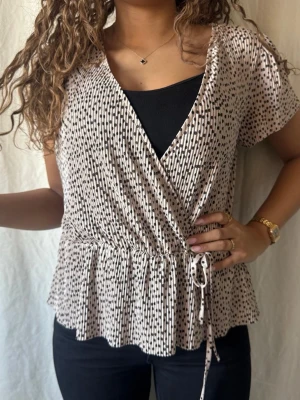 Beige omlottopp med knyt och mönster - Snygg prickig beige och svart omlottopp / peplumtopp med knyt i midjan. Toppen har korta ärmar, v-ringning och volang nedtill. Superfin nu till våren och sommaren med ett par jeans eller kjol. Pris kan diskuteras!🥰