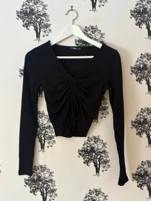 Svart ribbad croppad topp med snörning - Trendig svart croppad topp med lång ärm och v-ringning. Toppen har ribbad struktur och snygg snörning framtill som ger en cool rynkad effekt. Perfekt för dig som gillar stilrena och moderna plagg. Nopprig! 💓🌸