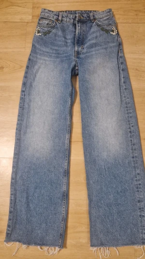 Vida blå jeans med broderi från Monki - Säljer ett par ljusblå jeans från Monki med breda ben och råa, fransiga benslut. Jeansen har coolt broderi i grönt och vitt vid fickorna fram och bak. Klassisk femficksmodell i denim med avslappnad passform, perfekta för en trendig streetstyle.