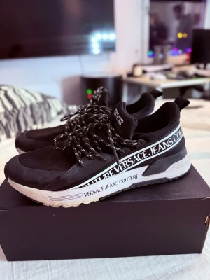 Svarta Versace Jeans Couture sneakers - Snygga svarta sneakers från Versace Jeans Couture med vit sula och tydlig logga längs sidan. Skorna har svarta och vita snören och en sportig design med låg profil. Materialet är en mix av textil och syntet. Perfekta för dig som gillar streetwear och exklusiva detaljer.