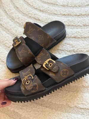 Louis Vuitton sandaler med monogram - Snygga Louis Vuitton sandaler i brunt konstläder med klassiskt monogrammönster och två justerbara remmar med guldfärgade spännen. Strl 37