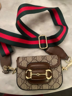 Gucci GG Supreme mini handväska - Säljer en Gucci GG Supreme mini handväska med klassiskt monogrammönster i beige och brunt, detaljer i brunt läder och guldfärgade metalldetaljer. Axelremmen är bred i grönt och rött med justerbart spänne. Perfekt för dig som gillar lyxiga accessoarer.