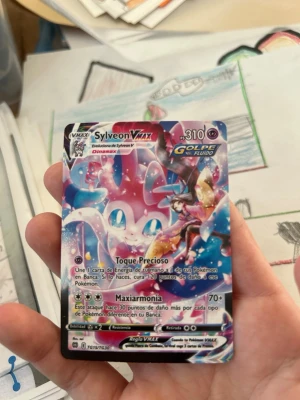Pokémonkort Sylveon VMAX Golpe Fluido - Säljer ett färgstarkt Pokémonkort med Sylveon VMAX från Golpe Fluido-serien. Kortet har en glansig yta och motiv i rosa, blått och vitt med anime-inspirerad design. Texten är på spanska och kortet är tillverkat i kartong. Perfekt för samlare eller Pokémonfans.
