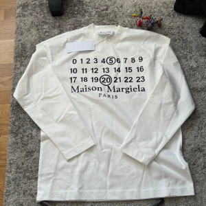  Maison Margiela longsleeve - Vit långärmad topp från Maison Margiela med tryckt logga och siffror på framsidan. Klassisk rak passform och rund halsringning. Tillverkad i mjuk bomull som känns skön mot huden. Perfekt för dig som gillar minimalistisk och stilren design.