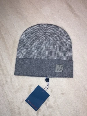 Grå Louis mössa med rutmönster - Snygg grå mössa med klassiskt rutmönster och uppvikt kant. Mössan har en diskret logga på en liten patch framtill. Perfekt accessoar för att lyfta din stil under kyliga dagar.