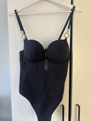 Svart body med gulddetaljer - Snygg svart body med vadderade kupor och dekorativa guldringar på axelbanden. Axelbanden är justerbara och baktill finns en smidig hyskknäppning. Materialet är stretchigt och följsamt för en bekväm passform. Liten cut-out framtill ger en extra detalj. Aldrig använt 