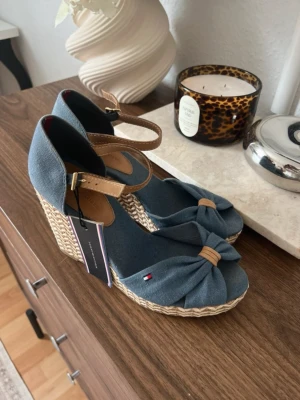 Blå kilklackssandaler Tommy Hilfiger (små i storlek)  - Snygga blå kilklackssandaler från Tommy Hilfiger med flätad sula och rosett framtill. Sandalerna har en justerbar rem runt ankeln i beige läderimitation och små logodetaljer på sidan. Perfekta för sommaren och ger en avslappnad men trendig look. (Är själv 38/39)