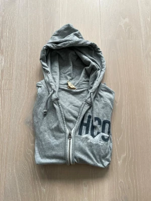 Grå zip hoodie från Hollister - Snygg grå hoodie från Hollister med dragkedja framtill och två fickor. Stor HCO-logga tryckt på bröstet och huva med dragsko. Tillverkad i mjukt bomullsmaterial, perfekt för chill dagar eller när du vill ha en avslappnad look.