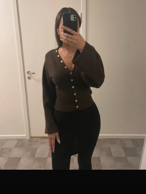 Brun ribbstickad kofta med knappar - Mörkbrun ribbstickad kofta med v-ringning och vita knappar framtill. Koftan har långa, vida ärmar och en åtsittande midja som ger en snygg siluett. Perfekt att styla med jeans eller kjol för en trendig look. (Inte mina bilder) 