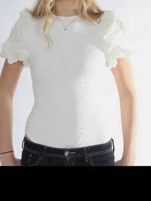 Vit body med puffärmar och blommor - Supersöt vit topp med puffiga kortärmar dekorerade med tygblommor. Tight passform och rund halsringning.