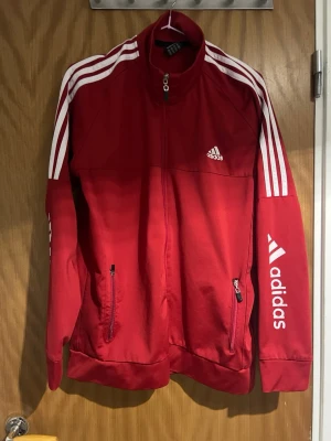 Röd Adidas träningsjacka - Snygg röd träningsjacka från Adidas med vita detaljer och klassiska tre ränder längs ärmarna. Jackan har dragkedja framtill, två fickor med dragkedja och Adidas-logga på bröstet och ärmen. Perfekt för träning eller chill. Materialet känns som polyester.