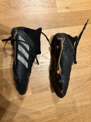 Adidas svarta fotbollsskor med socka stl 36 - Säljer ett par svarta Adidas fotbollsskor med klassiska vita ränder på sidan och bronsfärgade detaljer på sulan. Skorna har en strumpliknande hög krage för extra stöd runt ankeln och snörning för bra passform. Tillverkade i syntetmaterial och designade för spel på gräs.