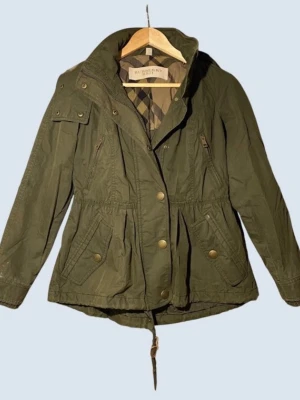 Olivgrön parkas från Burberry - Snygg olivgrön parkas från Burberry med klassiskt rutigt foder och luva med fuskpäls. Jackan har dragsko i midjan, flera fickor med knappar och detaljer i guld. Perfekt för kyliga dagar och ger en cool streetstyle-vibe.