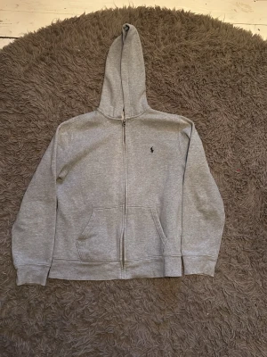 Grå zip hoodie från Ralph Lauren - Säljer en grå zip hoodie från Ralph Lauren med liten broderad logga på bröstet. Hoodien har dragkedja framtill, kängurufickor och en klassisk huva. Tillverkad i mjukt sweatshirtmaterial som är skönt att ha på sig.