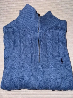 Blå kabelstickad tröja Polo Ralph Lauren - Kabelstickad blå tröja från Polo Ralph Lauren med halv dragkedja och hög krage. Tröjan har det klassiska broderade logotypen på bröstet och är gjord i mjukt garn. Perfekt för lager på lager och en chill look.