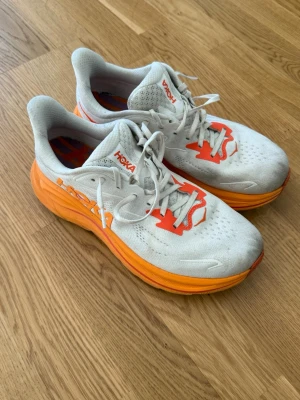 Hoka vita och orange löparskor - Säljer ett par snygga löparskor från Hoka i vitt med orange detaljer och tjock sula. Hoka Clifton 10, inköpta förra augusti så relativt nya och inte använda allt för mycket! Super sköna och snygga! Lite smutsiga då jag springer i skogen men det är ju bara att tvätta rent! 🤩💛🧡 säljer endast på grund av att jag fått ett par nya! Perfekta för dig som gillar att springa långt och vill ha bra dämpning Storlek 41 1/3 nypris: 1995kr..