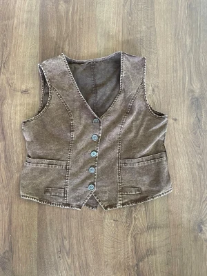 Brun vintageinspirerad väst med knappar - Snygg brun väst i tvättad bomull med fem dekorativa metallknappar framtill och två fickor. Västen har markerade sömmar och en cool, retroinspirerad look. Perfekt att styla över en t-shirt eller skjorta för en trendig lager-på-lager känsla.