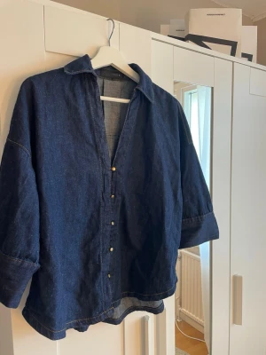 Mörkblå denimskjorta från Zara - Säljer en mörkblå denimskjorta från zara med klassisk krage och knappar framtill. Skjortan har en loose passform och är tillverkad i bomull. Perfekt för dig som gillar en avslappnad och cool stil.