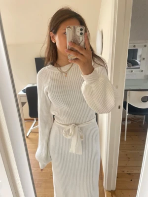 Vit ribbad långklänning med knyt - Säljer en vit ribbad långklänning med knyt i midjan och långa ärmar. Klänningen har en snygg v-ringning och är stickad, vilket gör den både bekväm och trendig. Perfekt för dig som gillar en clean och stilren look. Är 168 cm lång. 