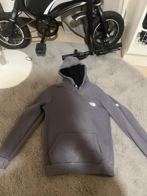 Grå hoodie från The North Face - Säljer en grå hoodie från The North Face med klassisk logga på bröstet. Tröjan har en stor huva med svart innerfoder och en praktisk magficka. Perfekt för dig som gillar streetwear och vill ha något bekvämt och stilrent.