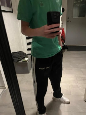 Palm Angels svart mjukisbyxa - Svarta mjukisbyxor från Palm Angels med vita sidoremsor och logotyp på låret. Byxorna har en avslappnad passform och resår i midjan. Perfekta för chill dagar och streetwear-stil.