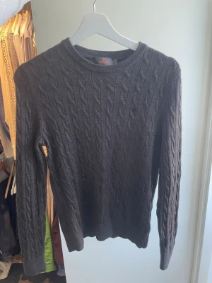 Brun kabelstickad tröja Morris - Snygg brun kabelstickad tröja från Morris med rund halsringning och diskret logga på bröstet. Tröjan har långa ärmar och ribbade muddar vid ärmslut och nederkant. Perfekt för dig som gillar klassisk och stilren design.