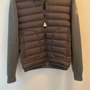 Grå Moncler pufferjacka med dragkedja - Snygg grå pufferjacka från Moncler med quiltad front och mjuka stickade ärmar. Jackan har två fickor med dragkedja och en cool Moncler-logga på ärmen. Perfekt för dig som vill ha en stilren och sportig look under höst och vinter.