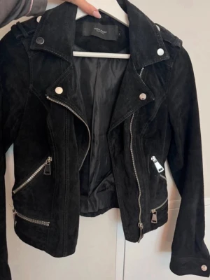Svart skinnjacka från Vero Moda - Cool svart mocka jacka från Vero Moda med klassisk bikerstil, silverfärgade dragkedjor och knappar. Jackan har snedställd dragkedja framtill, flera fickor med zip och detaljer på axlarna. Perfekt för att lyfta din outfit.