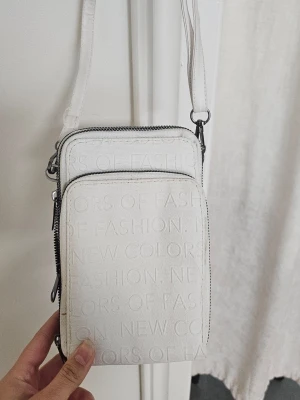 Vit axelväska med präglad text - Säljer en stilren vit axelväska med präglad text över hela väskan. Väskan har två fack med dragkedja och justerbar axelrem. Materialet känns som konstläder och designen är minimalistisk men ändå trendig.