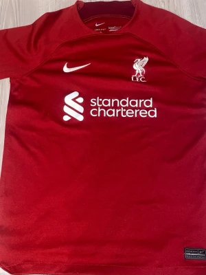 Liverpool matchtröja Nike röd - Säljer en röd Liverpool FC matchtröja från Nike med klubbmärke och sponsortryck framtill. Tröjan har korta ärmar och är tillverkad i ett lätt och ventilerande material, perfekt för fotboll eller träning. Storlek 147-158