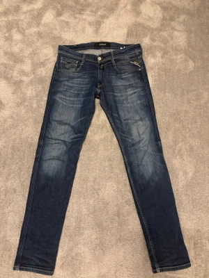 Blå Replay jeans med snygga detaljer - Säljer ett par blå jeans från Replay med klassisk femficksdesign och snygga slitningar framtill. Jeansen har raka ben, kontrastsömmar och läderpatch med logga bak i midjan. Perfekta för en avslappnad och trendig look.