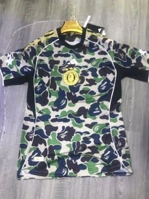 Camouflage topp från Adidas x Bape - Unik kortärmad topp från Adidas x Bape med grönt, svart och blått camouflage-mönster. T-shirten har rund hals, vita detaljer och ett gult emblem på bröstet. Perfekt för dig som gillar streetwear och vill sticka ut.