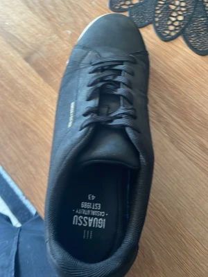 Svarta sneakers från Jousasu - Stilrena svarta sneakers från Jousasu med vit sula och klassisk låg profil. Skorna har snörning och ovandel i mockaliknande material. Märkeslogga syns på sidan och innersulan. Perfekta för en clean och enkel look.