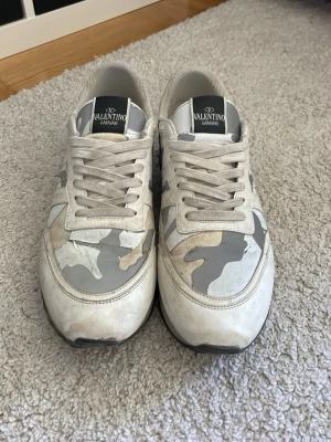 Valentino camo sneakers i vitt och grått - Snygga sneakers från Valentino Garavani med grå och vita camo-detaljer. Skorna har lågt skaft, vita snören och en mix av läder och textil i ovandelen. Perfekta för dig som vill sticka ut med exklusiva och trendiga sneakers.