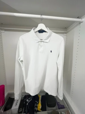 Vit långärmad piké från Polo Ralph Lauren - Klassisk vit långärmad pikétröja från Polo Ralph Lauren med krage, knappar framtill och den ikoniska broderade loggan på bröstet. Tillverkad i bra kvalitet som även känns skön mot huden. Perfekt till jeans eller chinos för en clean look.