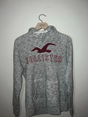 Hollister hoodie - Gråmelerad hoodie från Hollister med klassisk logga i vinrött. Mjuk och bekväm med avslappnad passform, perfekt för vardag. Stilren, lättmatchad och tidlös – ett säkert kort i garderoben. Fint skick bortsett från en liten gul fläck syns på bild 4. Priset kan diskuteras vid snabb affär!